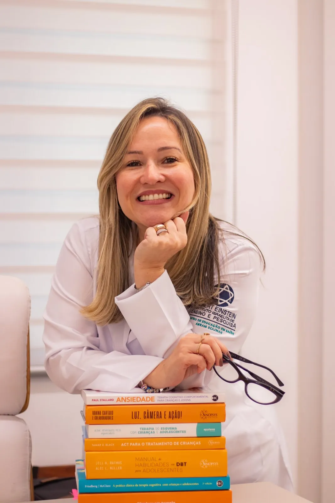 Psi. Raquel Peyroton sorrindo, de jaleco, apoiada sobre livros